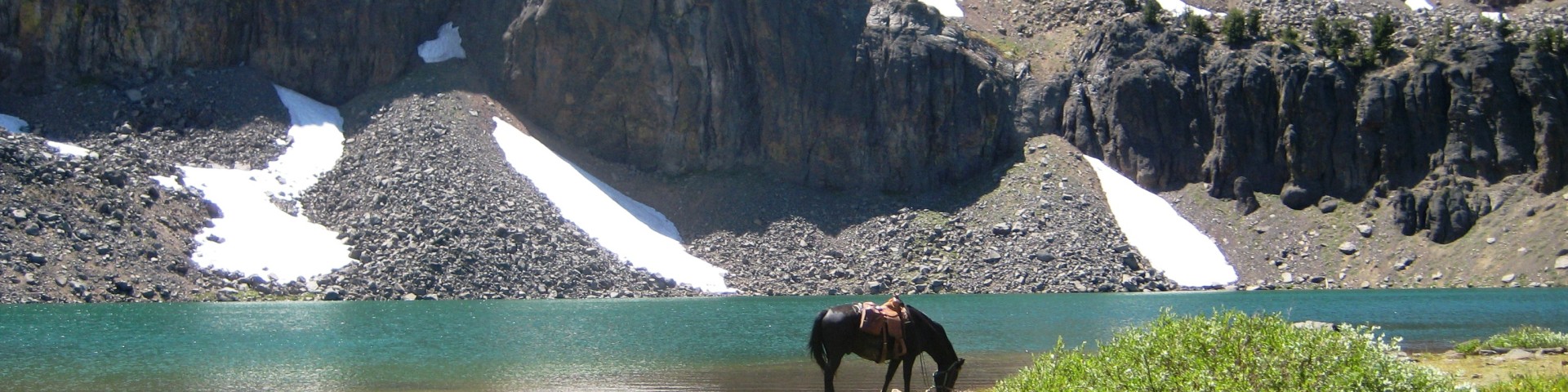 A tranquil mountain lake below steep volcanic cliffs in the South Warner Wilderness. A horse stands at the water's edge drinking, with patches of lingering summer snow scattered across the rocky slopes behind it. Bright turquoise water, rugged ridgelines, and sparse trees create a remote, high-elevation alpine scene.